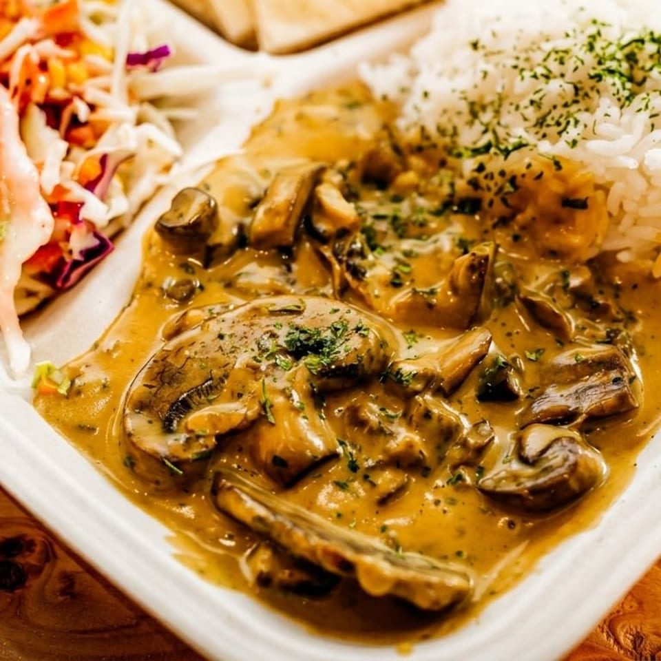Takeaway tray with mushrooms in a creamy sauce, white rice, and colorful slaw on a wooden table.